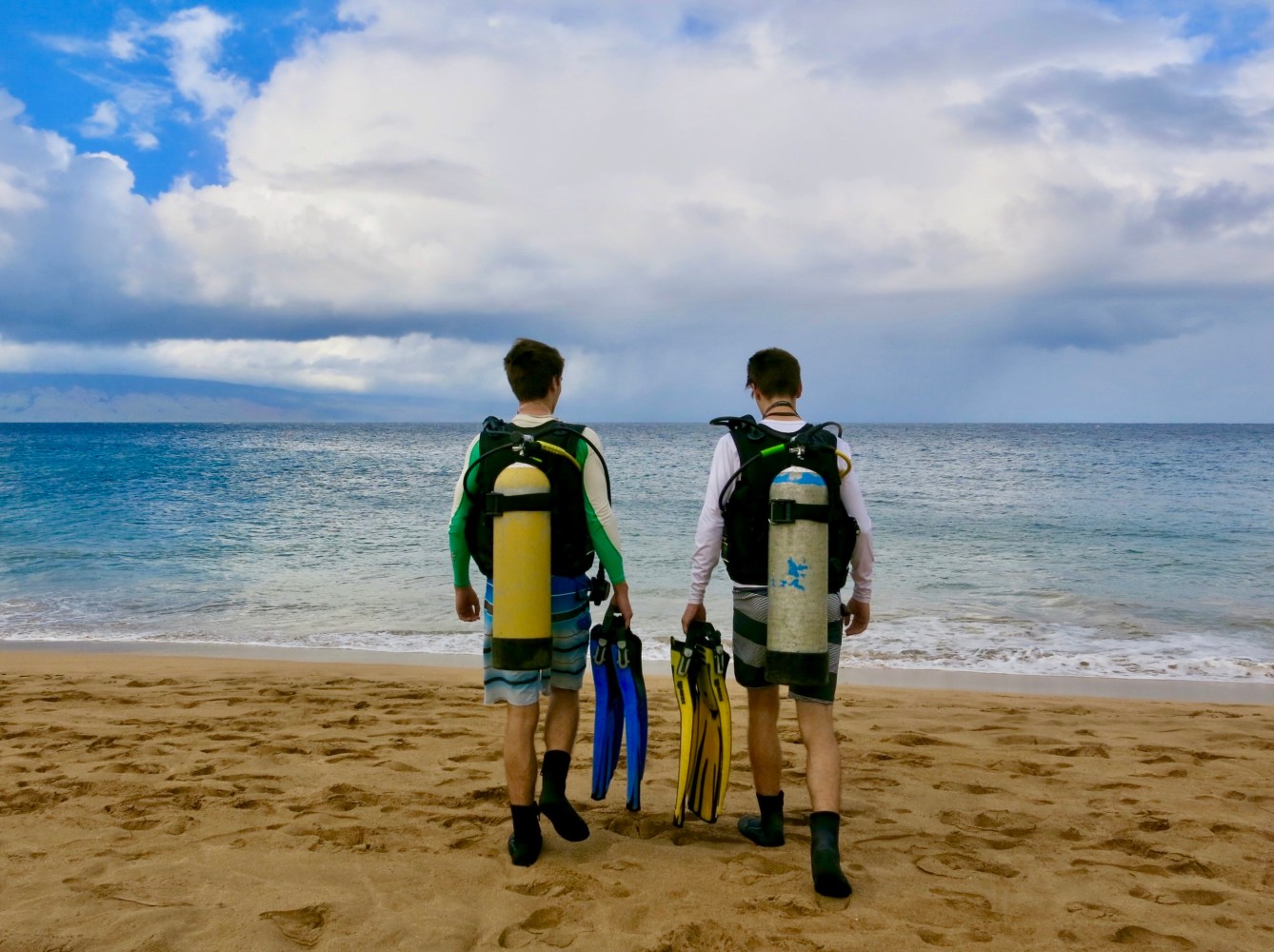 Two scuba divers wearing tanks and carrying fins walk across a sandy Maui beach toward the ocean.
