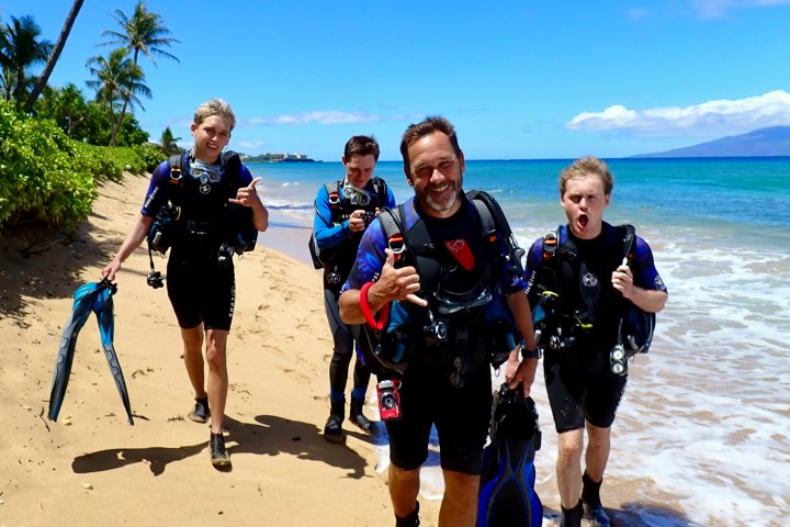 Four scuba divers in dive gear standing on a sandy Maui beach with turquoise water and palm trees behind them.